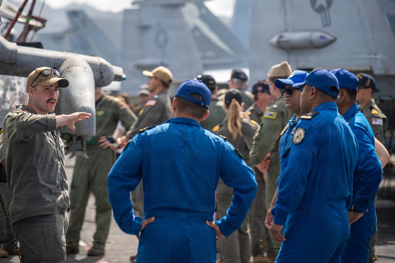 A man in a brown military uniform gestures near a group of four men dressed in blue flight suits while standing on the flight deck of a Navy aircraft carrier.