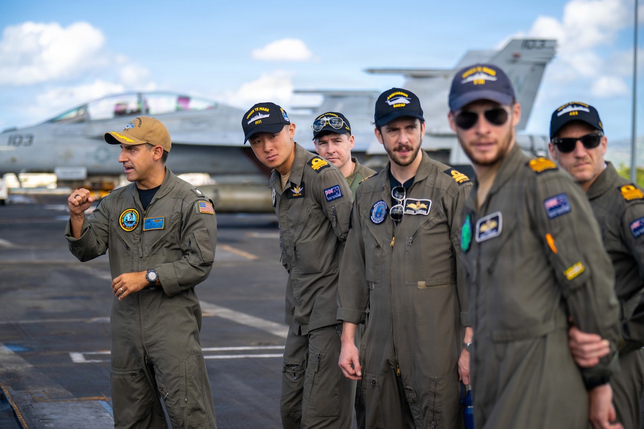A group of men in military flight suits look to their left down the flight line of an aircraft carrier while another man in similar attire is looking to the right.