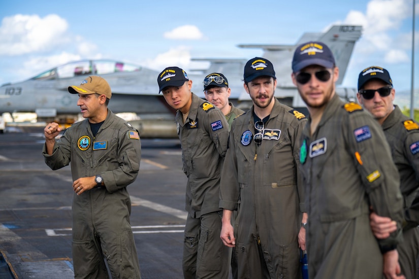 A group of men in military flight suits look to their left down the flight line of an aircraft carrier while another man in similar attire is looking to the right.