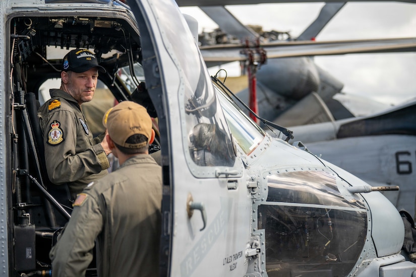 :  Two men in military flight suits talk to each other while one of them is sitting in the cockpit of a military helicopter.
