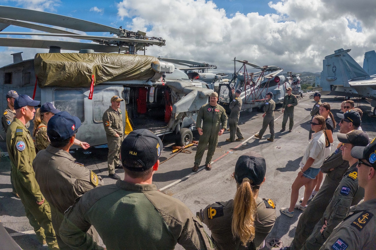 Two men in military flight suits stand in front of a military helicopter as a large group of people wearing flight suits and a couple of people in civilian attire look on.
