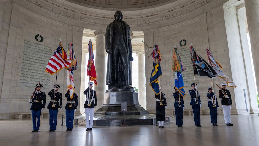 Nine people in formal military uniforms from each military service branch hold flags beside a statue in a rotunda.