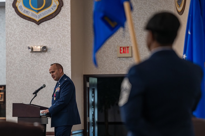 A Colonel is speaking at a podium while an individual closer to the camera is blurred, but you can closely make out a Command Chief rank insignia.
