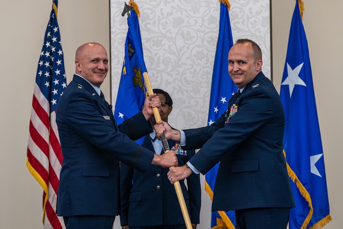 3 individuals stand on stage. The one on the left is is being given the guidon by the one on the right.