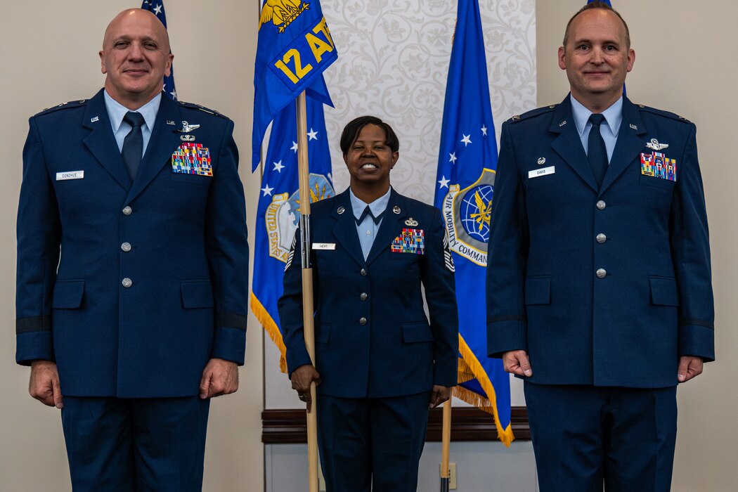3 individuals in dress blues stand at attention in front of flags. The one in the middle is holding a guidon.