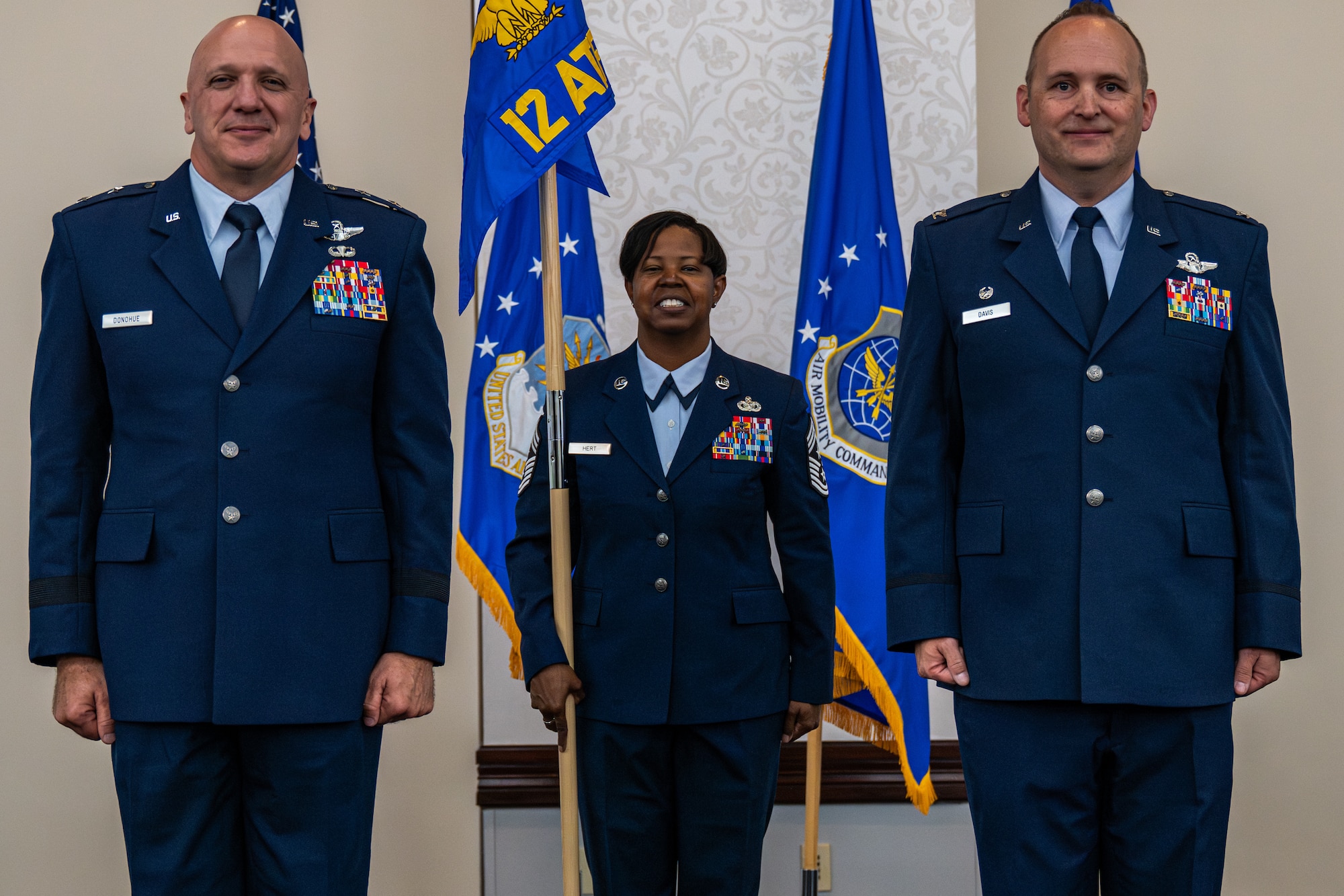 3 individuals in dress blues stand at attention in front of flags. The one in the middle is holding a guidon.