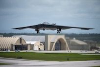 A stealth bomber takes off from an airfield with aircraft hangars behind it