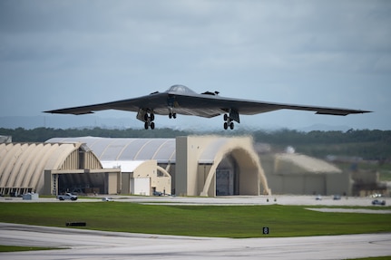 A stealth bomber takes off from an airfield with aircraft hangars behind it