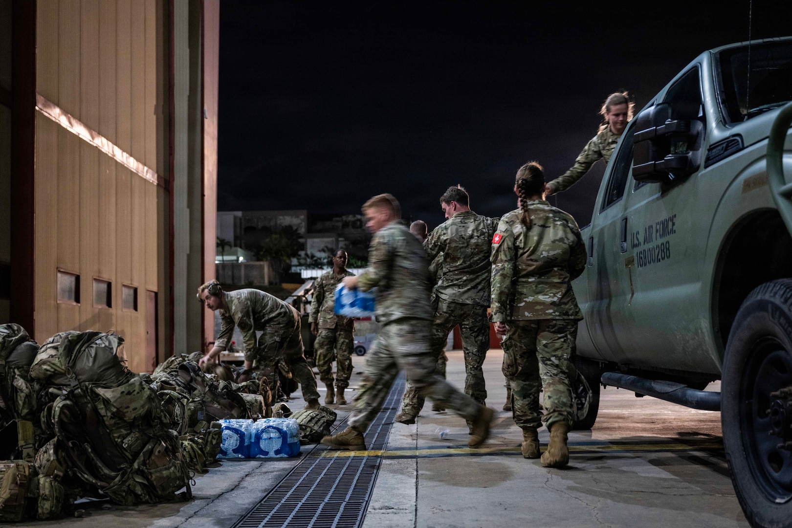 Airmen in camouflage attire unload packages of water bottles from a pickup truck as their backpacks rest against the wall of a building.