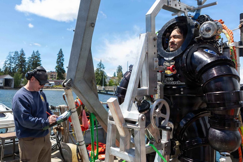 A man wearing casual attire marks a checklist while standing on a boat as another man stands in a dive suit.