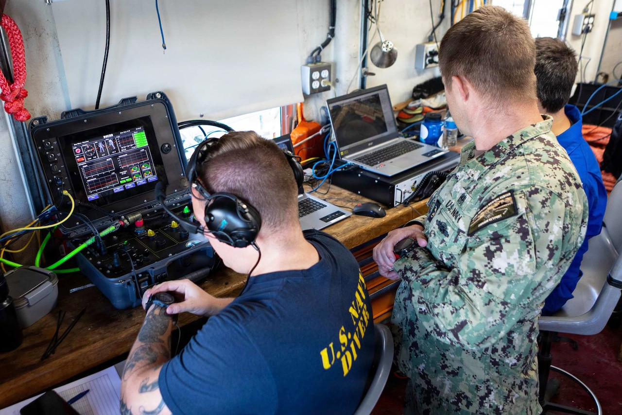 A man wearing a camouflage military uniform stands in a room looking at a computer while two other people in casual attire sit while checking equipment.