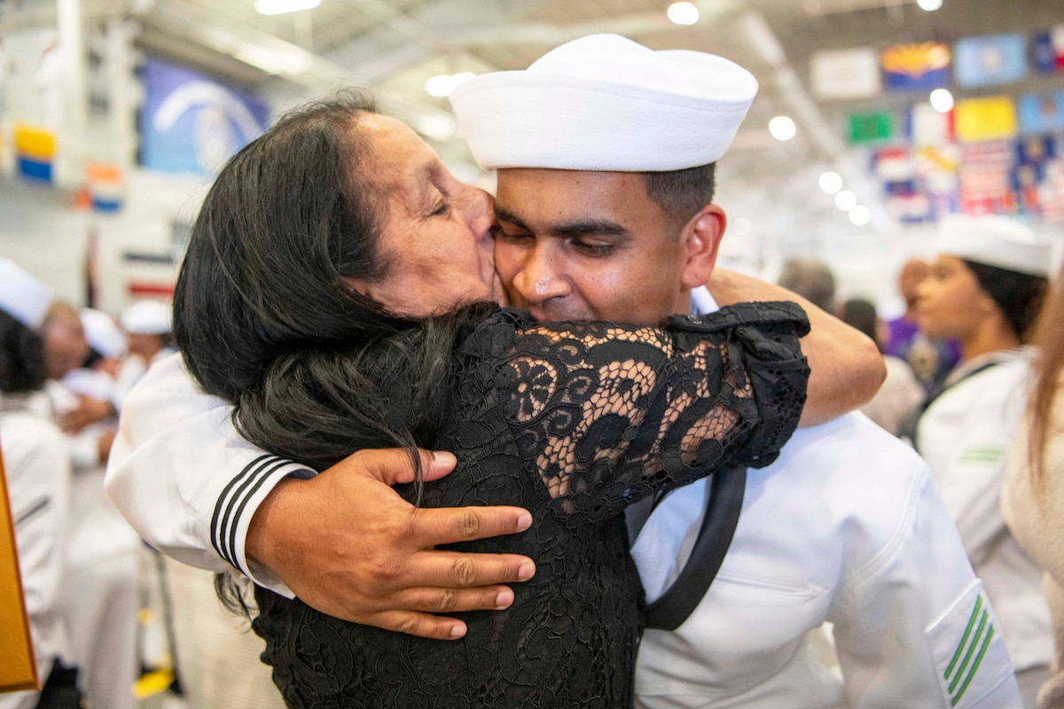 A loved one in a black dress kisses a sailor in a white dress uniform on the cheek while other sailors in similar uniforms are seen in a blurred background.