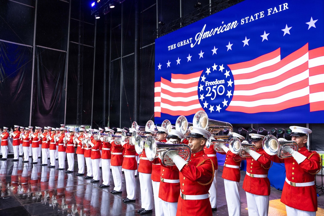 Marines in red dress uniforms play instruments in formation on black stage with a blue, red and white banner in the background.