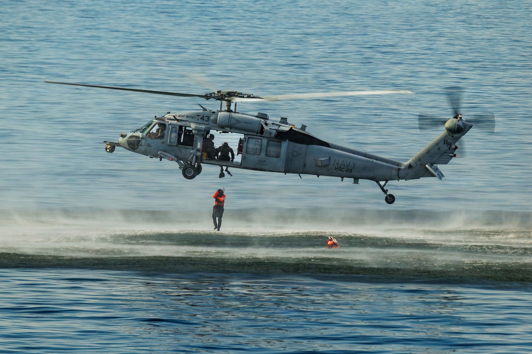 A sailor wearing a life vest jumps from a hovering helicopter into a body of water.