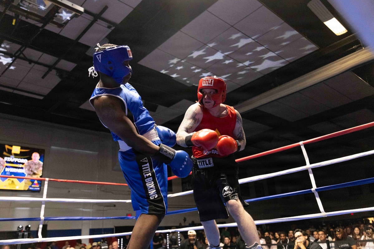 Two soldiers, one in blue fighting gear the other in red, box in a dark ring with stars on the ceiling as a seated audience watches.
