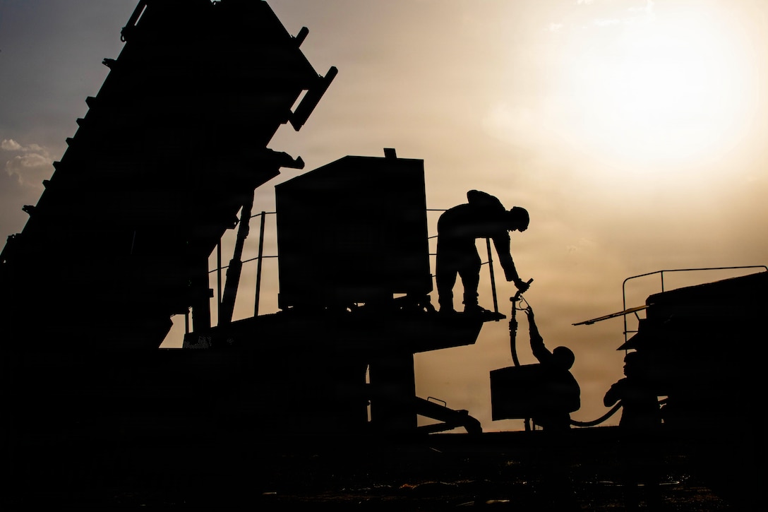 A soldier reaches down from a deck to grab a fuel line that another soldier is handing to the soldier. They are in silhouette with the sun behind clouds in the background.