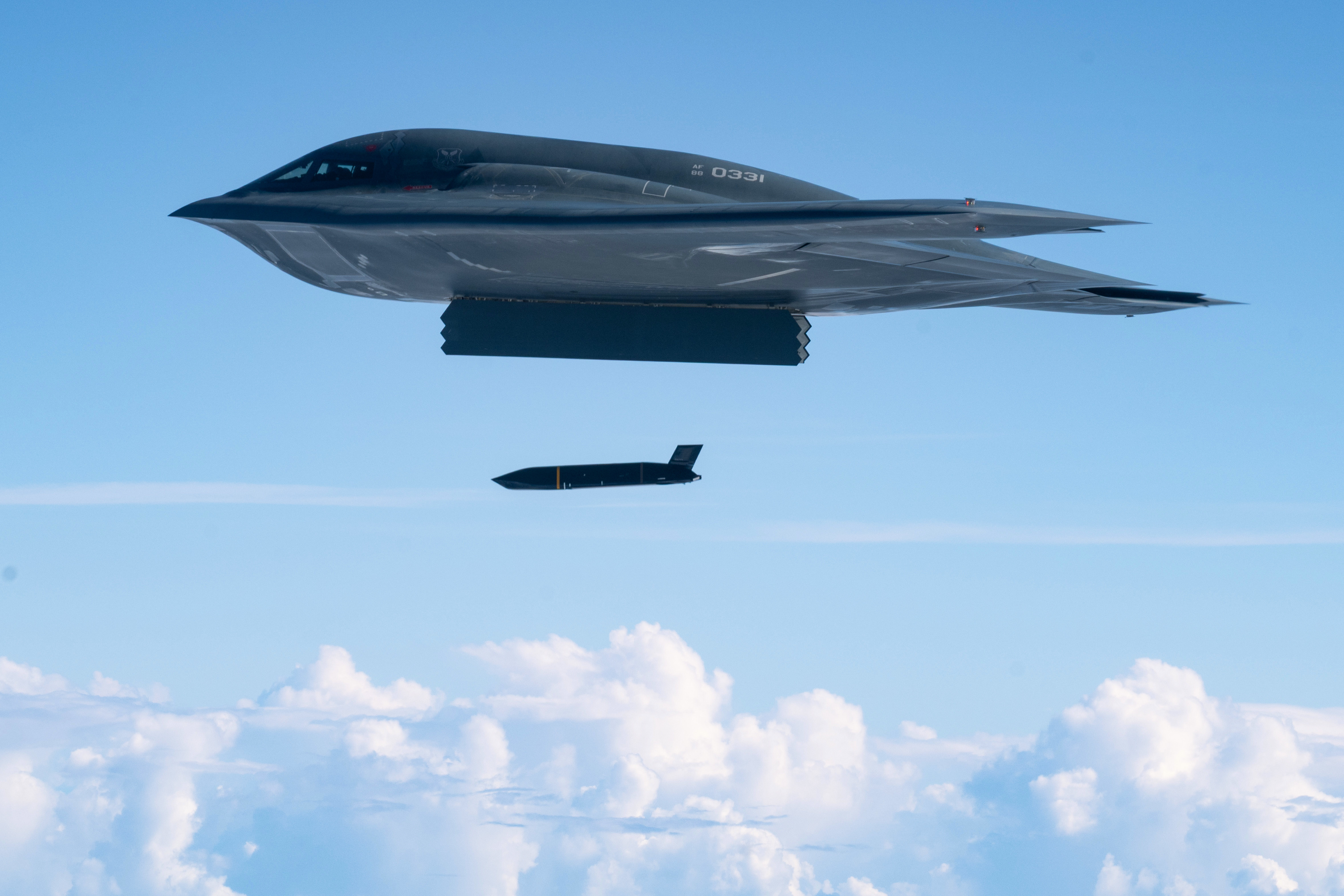 A B-2 aircraft drops a missile while flying over puffy clouds in a light blue sky.