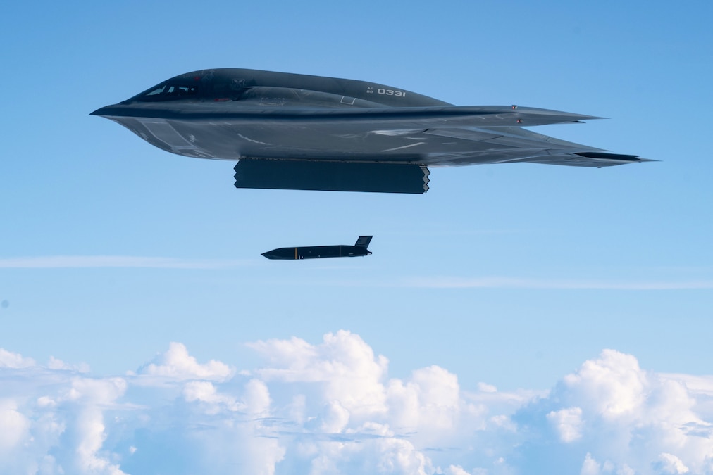 A missile launches from a stealth bomber in the air, with clouds below it and a light blue sky in the background.