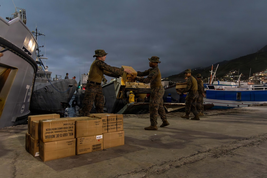 Service members in camouflage attire handle boxes at a port, with a gray sky and boats in the background.
