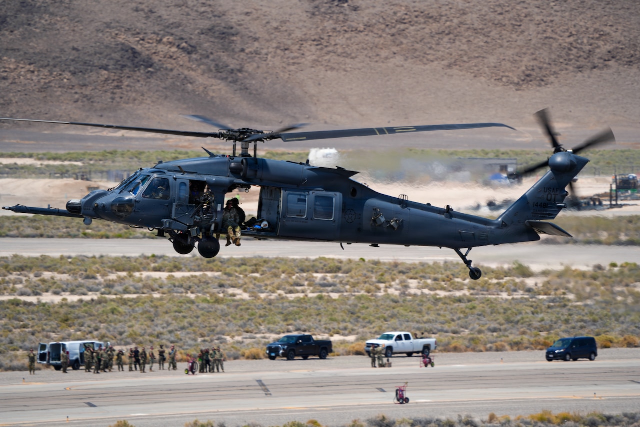 A military helicopter hovers above a flight line as people in military camouflage uniforms stand around on the ground.