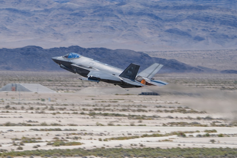A military jet flies over desert terrain.