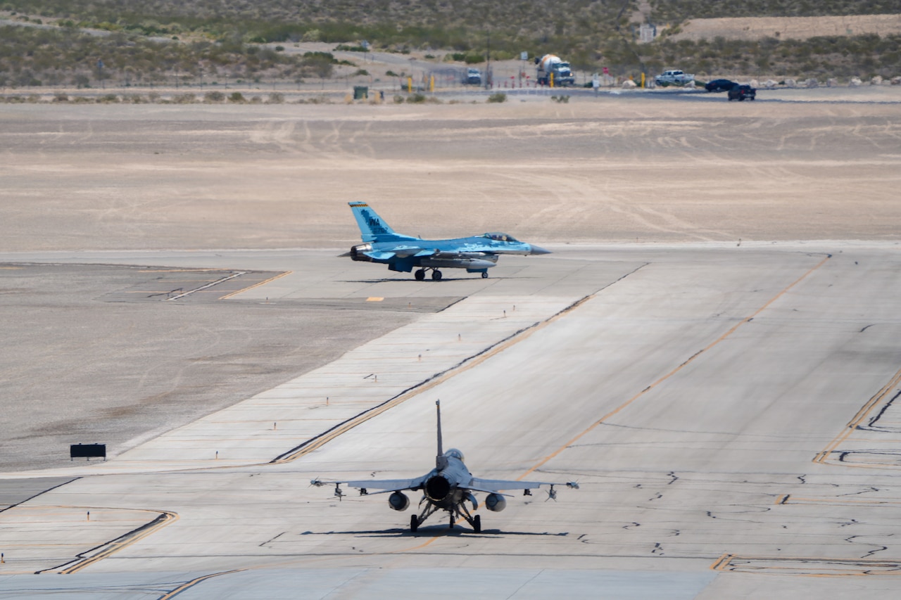 Two military jets taxi on a flight line.