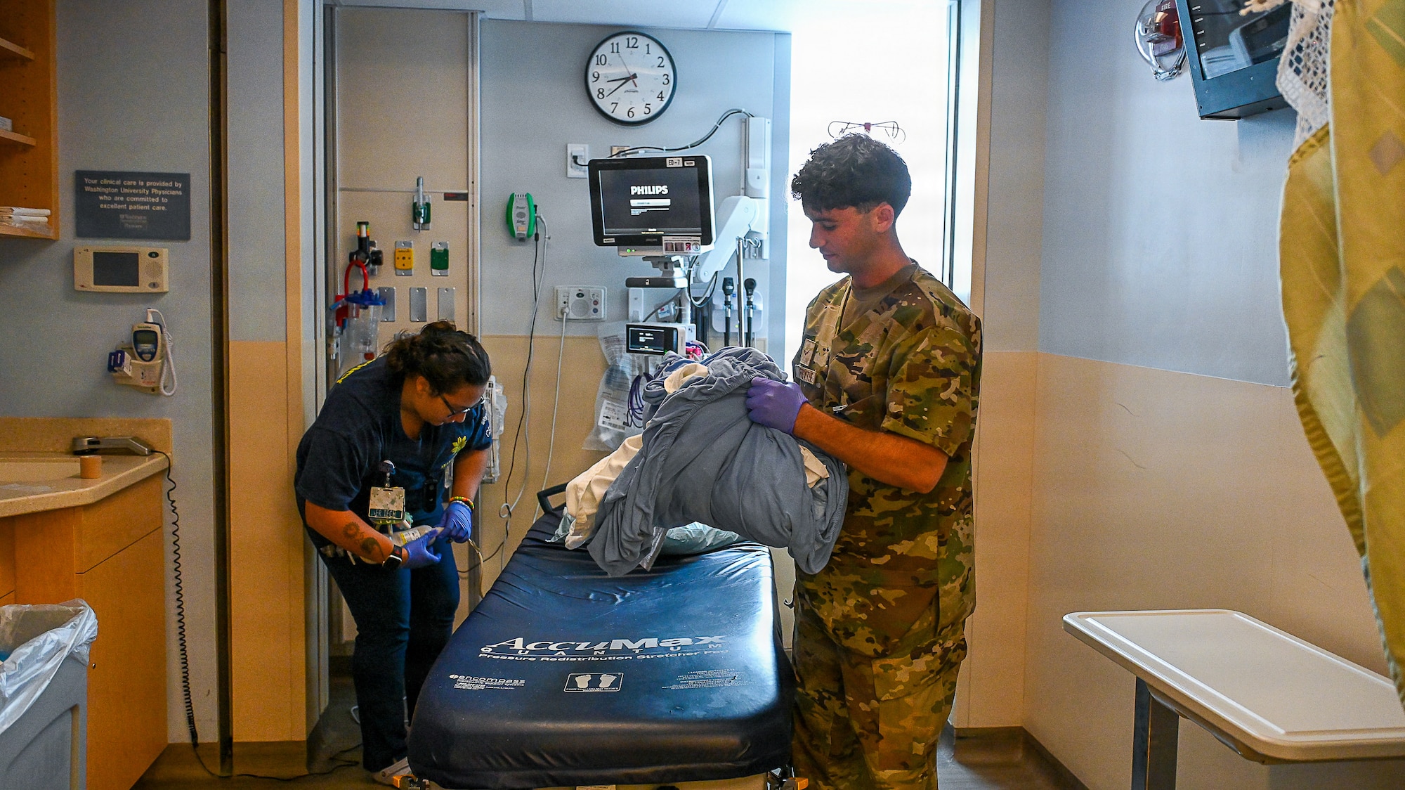 2 medical professionals remove bedding from a gurney in an emergency room.