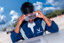 A photo of a woman wearing vision-impairing goggles outside attempting to throw a tennis ball into a bucket.