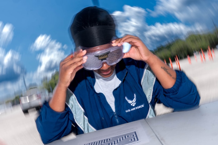 A photo of a woman wearing vision-impairing goggles outside attempting to throw a tennis ball into a bucket.