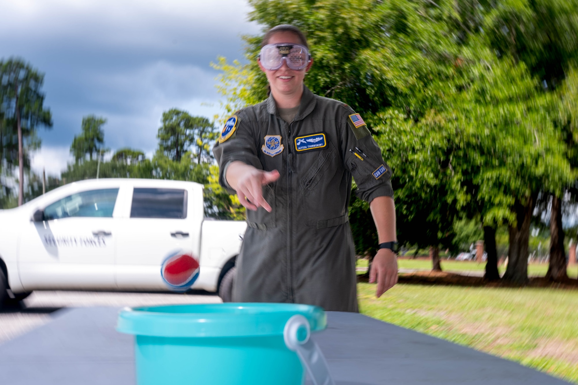 A photo of a woman wearing vision-impairing goggles outside attempting to throw a tennis ball into a bucket.