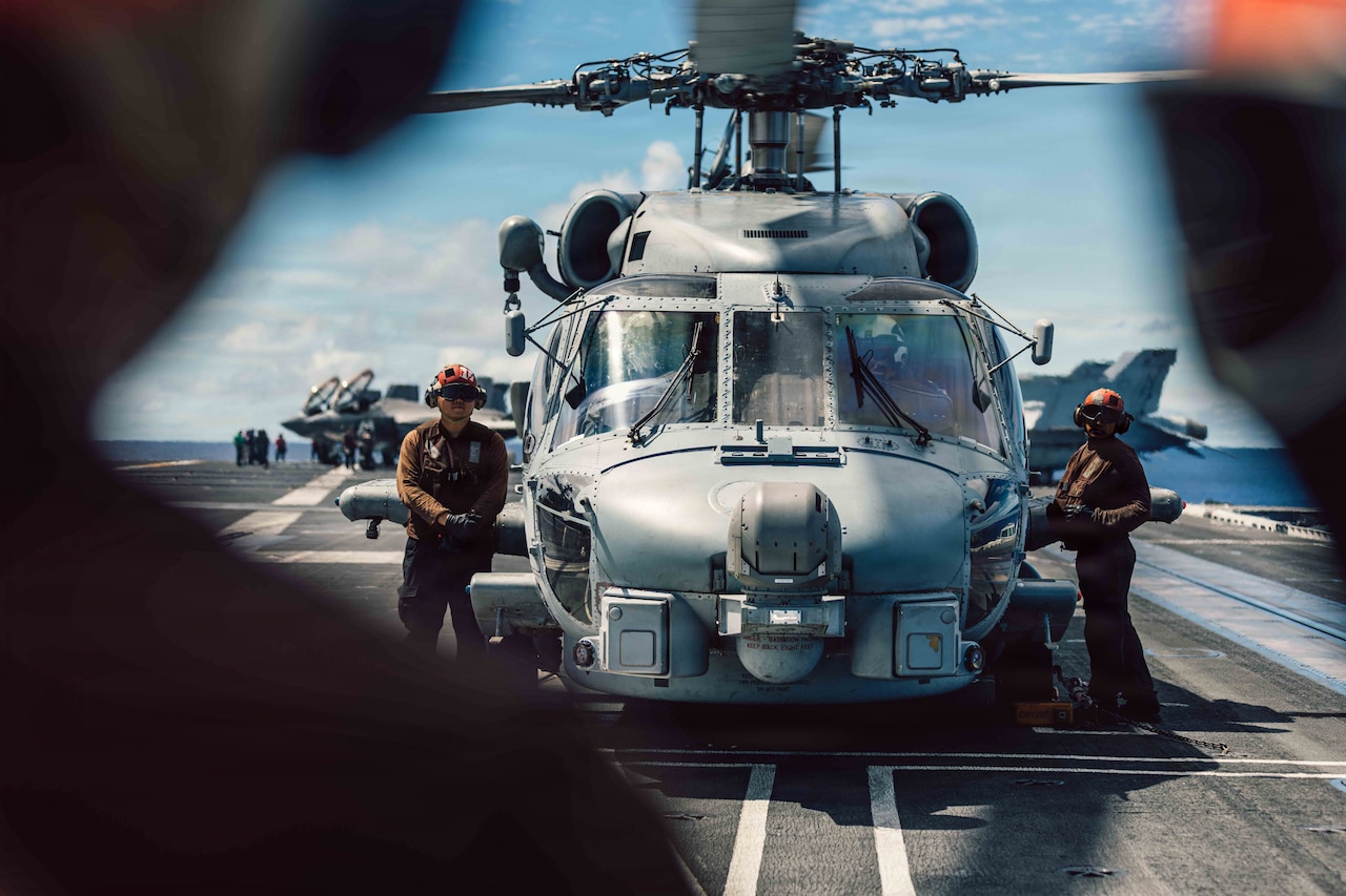 Two people in military uniforms stand next to a military helicopter on the flight deck of an aircraft carrier as a person in the foreground signals them.