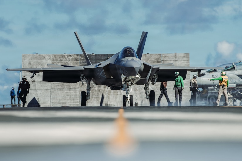 Several people in military uniforms walk around a military aircraft on the flight deck of an aircraft carrier.
