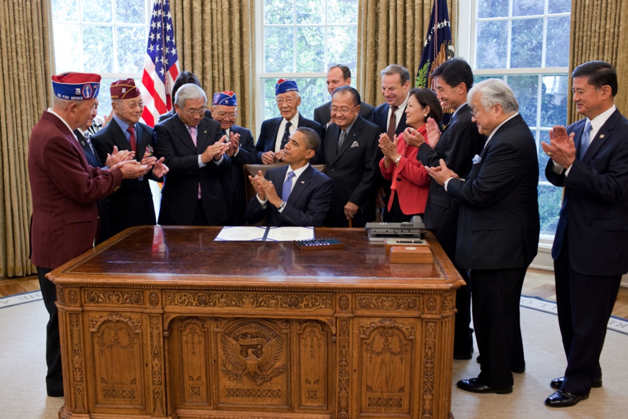 A man sits at a desk while people around him are smiling and clapping.