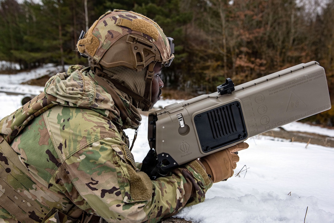 A soldier points a rectangular tan-colored weapon while positioned low in the snow.