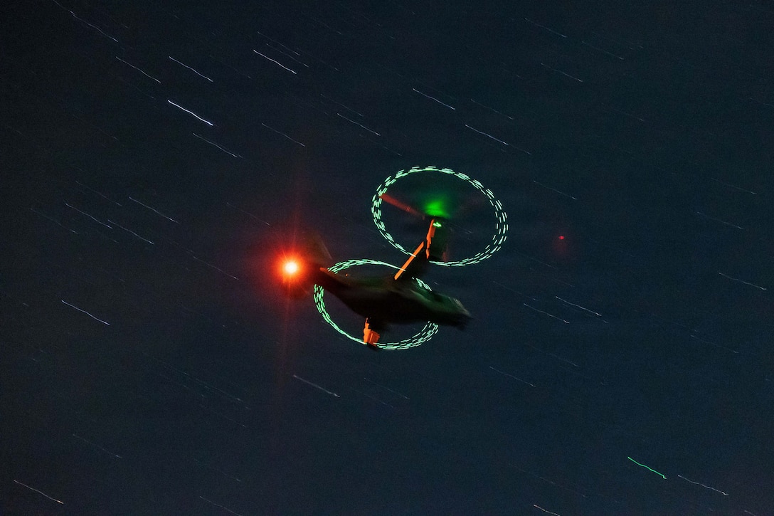 The prop rotors on an osprey aircraft creates two green circles as it flies against a moving dark sky.