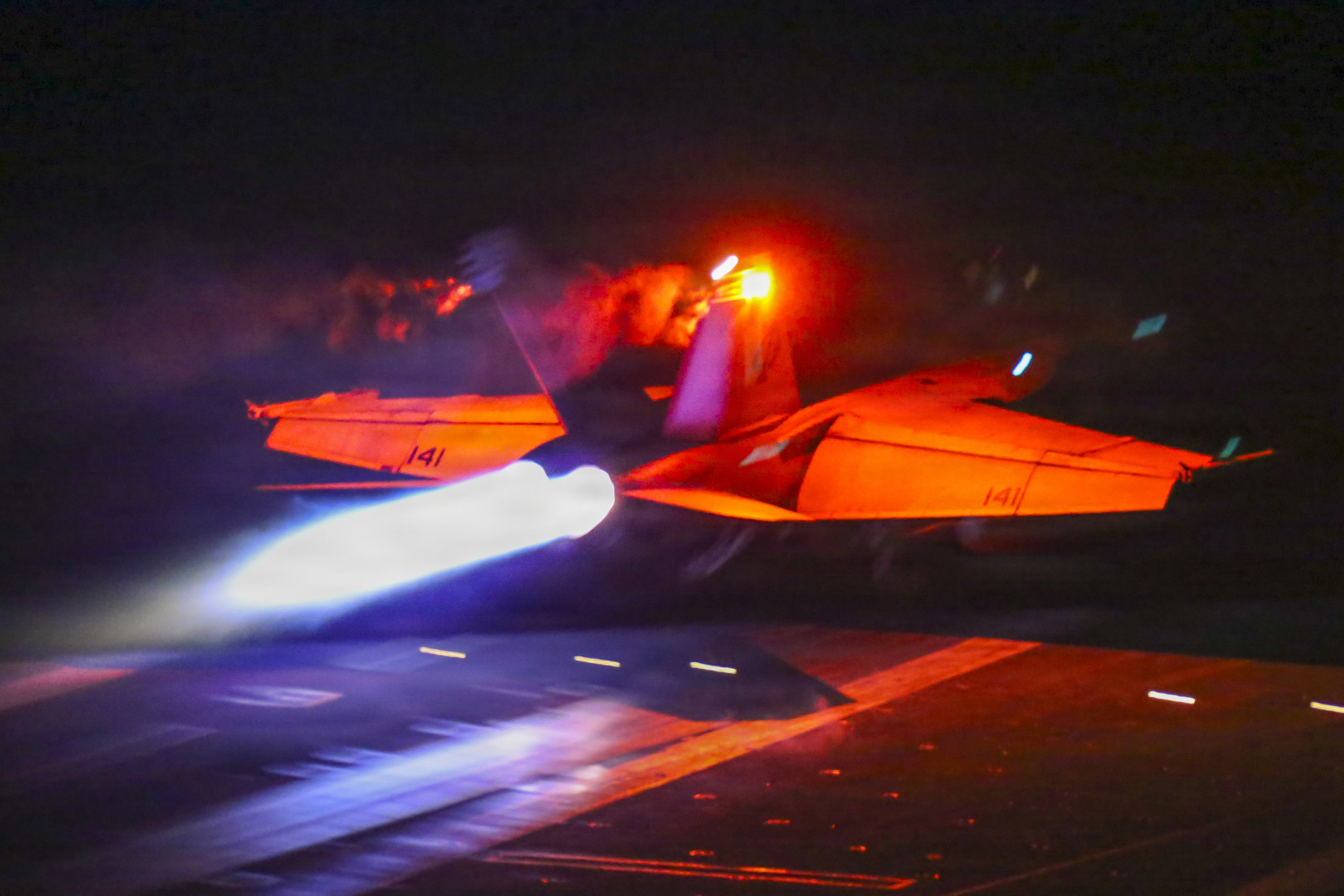 A jet lit with red-orange light takes off in dark conditions from the deck of a ship, with light blue flame shooting from the back.