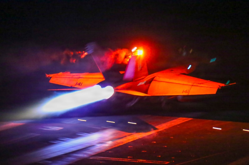 A jet lit with red-orange light takes off in dark conditions from the deck of a ship, with light blue flame shooting from the back.