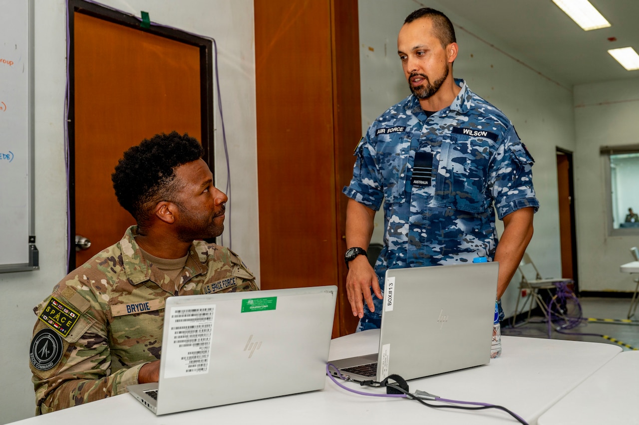 Two men, one seated and wearing a green camouflage military uniform and another standing and wearing a blue camouflage uniform, look at each other. There is a table with laptops in front of them.