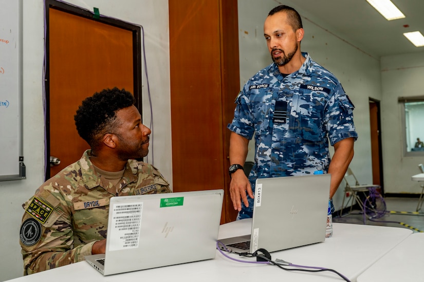 Two men, one seated and wearing a green camouflage military uniform and another standing and wearing a blue camouflage uniform, look at each other. There is a table with laptops in front of them.