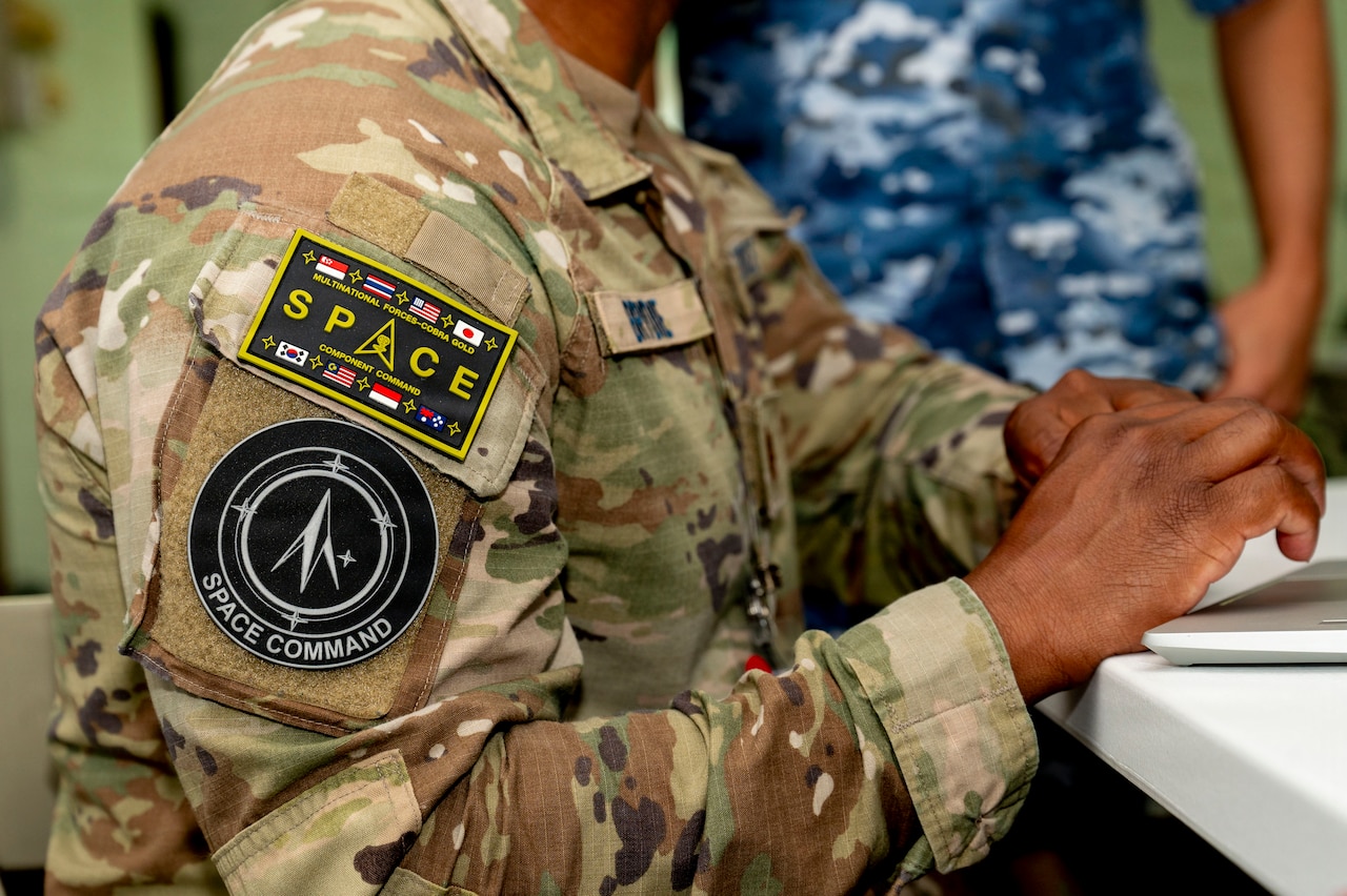 A Space Force patch is shown on the shoulder of a person wearing a camouflage military uniform. Another person in similar attire is standing in the background.