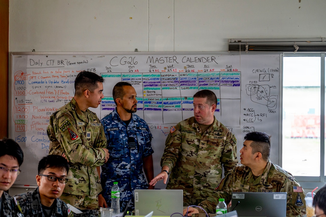 Three men wearing camouflage military uniforms from multiple nations stand in front of a white board. There are other men in similar attire seated at tables with laptops in front of them.