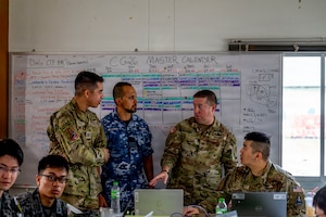 Three men wearing camouflage military uniforms from multiple nations stand in front of a white board. There are other men in similar attire seated at tables with laptops in front of them.
