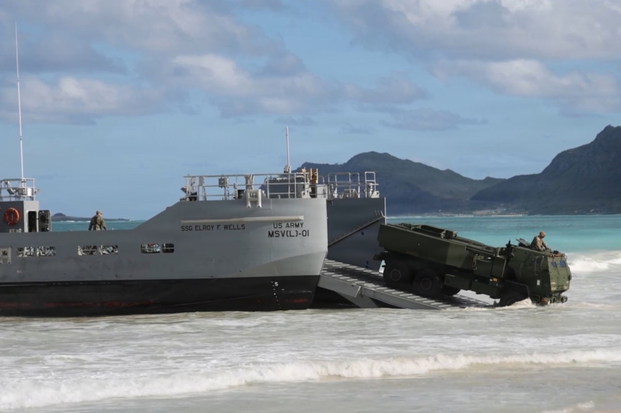 A person in a camouflage military uniform drives a large vehicle down the ramp of a ship onto a beach. There are mountains and blue-green water in the background.