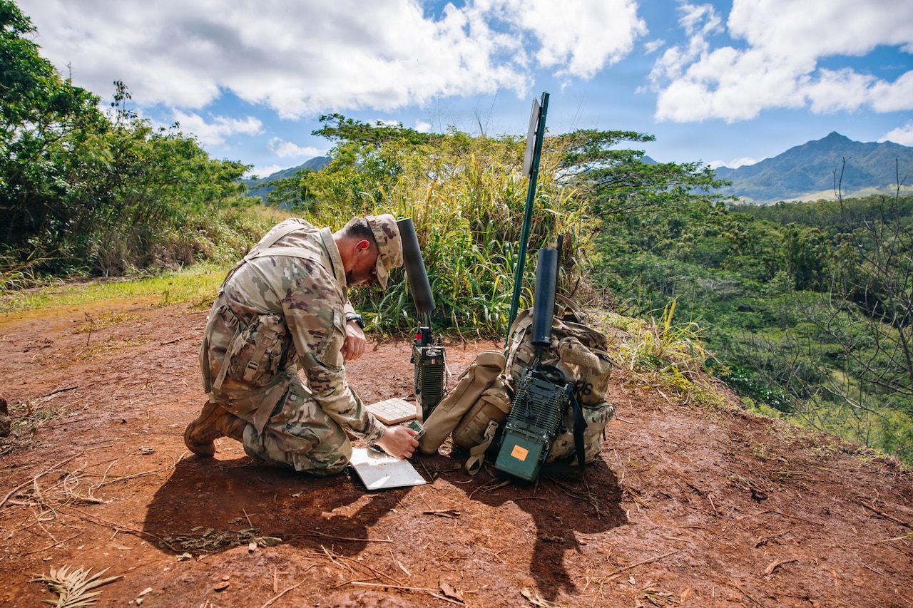 A man in a camouflage military uniform kneels on a hill in the forest while looking at a tablet. Military communication equipment is gathered next to him.