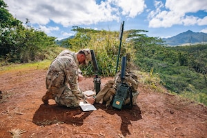 A man in a camouflage military uniform kneels on a hill in the forest while looking at a tablet. Military communication equipment is gathered next to him.