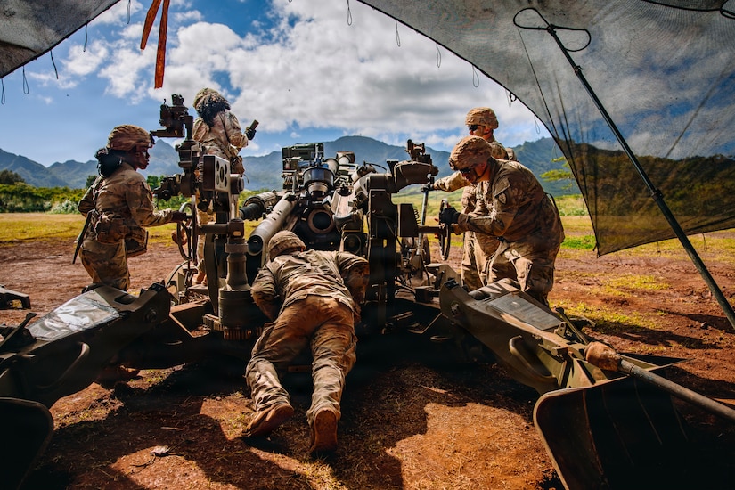 Five people in camouflage military uniforms stand under a tent and prepare to fire a large military weapon in a field, with mountains in the distance.