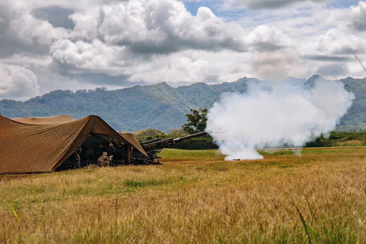 Several people in camouflage military uniforms stand in a tent while firing a large military weapon in an open field. The sky is cloudy, and there are mountains in the background.