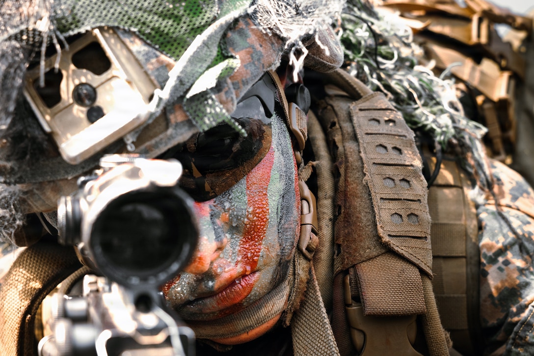 A Marine in camouflage face paint looks through a weapon scope.