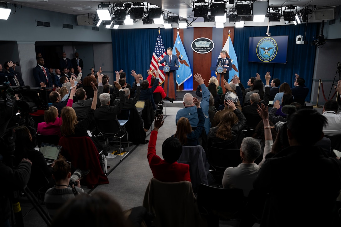 A man in business attire and a man in a formal military uniform stand behind lecterns on a stage in a large room and speak to the crowd before them. Behind them are an American flag, two flags with eagles in the center and a sign on the wall that reads, The Pentagon, Washington.