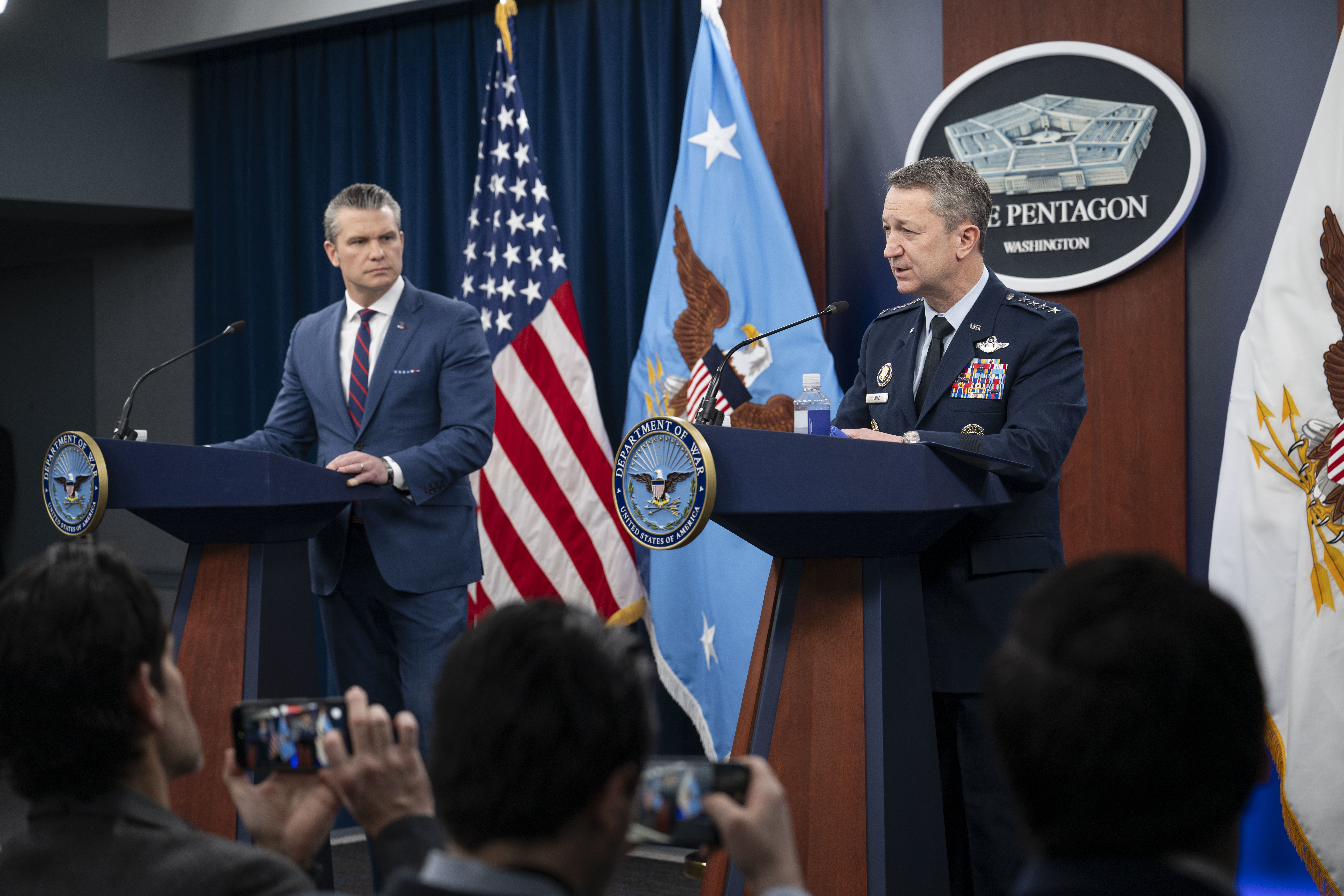 A man in business attire and a man in a formal military uniform stand behind lecterns and speak to the crowd before them. Behind them are an American flag, two flags with eagles and a sign that reads, The Pentagon, Washington.